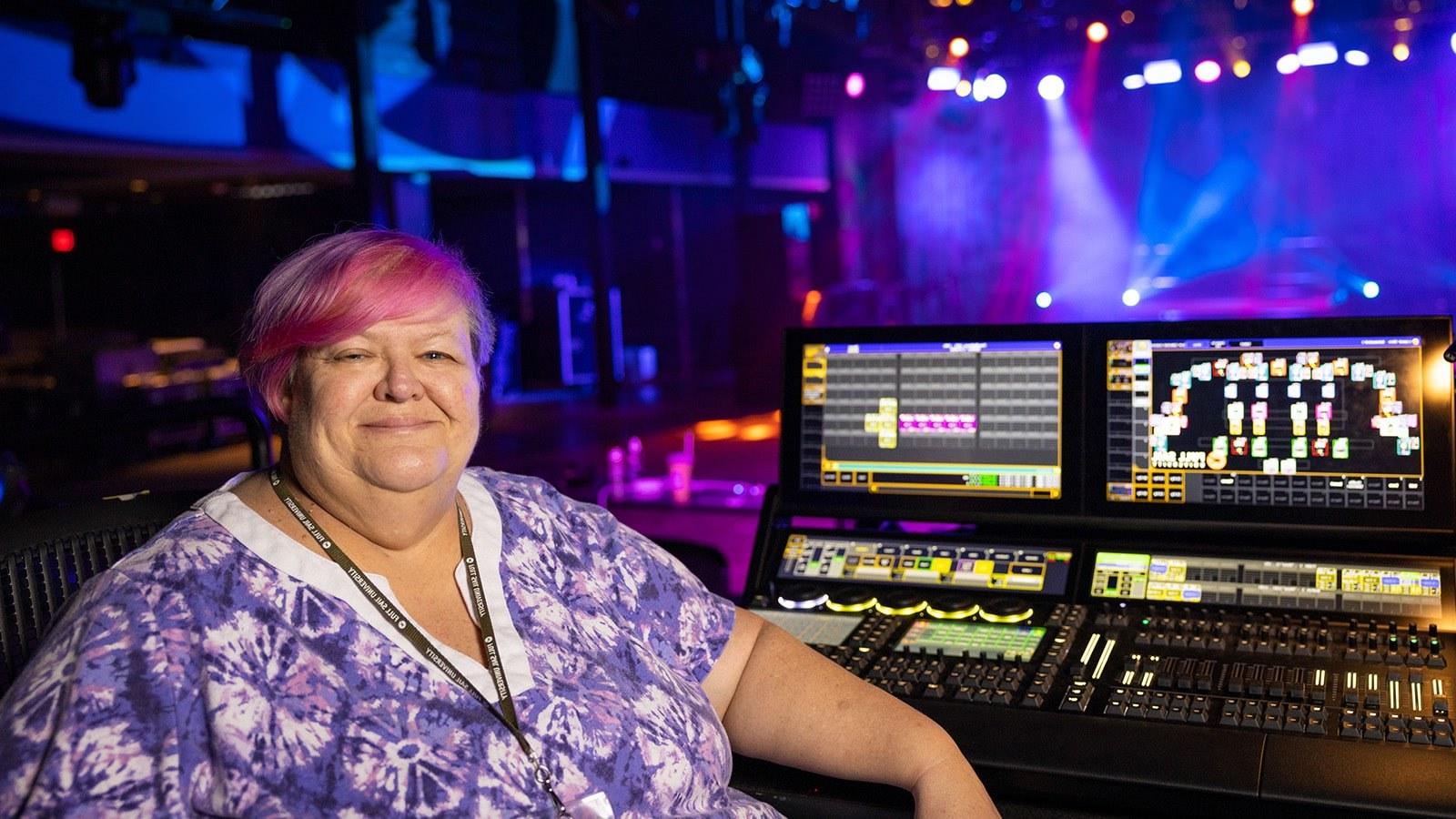 Susan Kelleher sits at a lighting console. 她身后的舞台被紫色、蓝色和白色的灯光照亮.